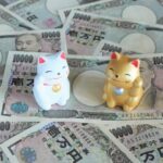 ばらまかれた一万円札の上に置かれた広い招き猫と金色の招き猫